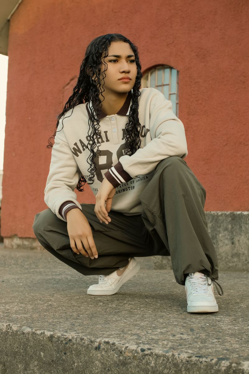A young adult squatting in trendy streetwear against a warm-toned urban wall.